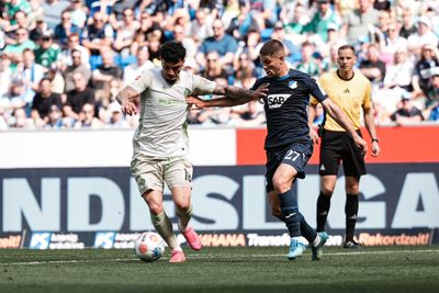 Zwei Fußballspieler, einer in Weiß und einer in Blau, kämpfen während eines Spiels um den Ball, im Hintergrund sind ein Schiedsrichter und Zuschauer zu sehen.