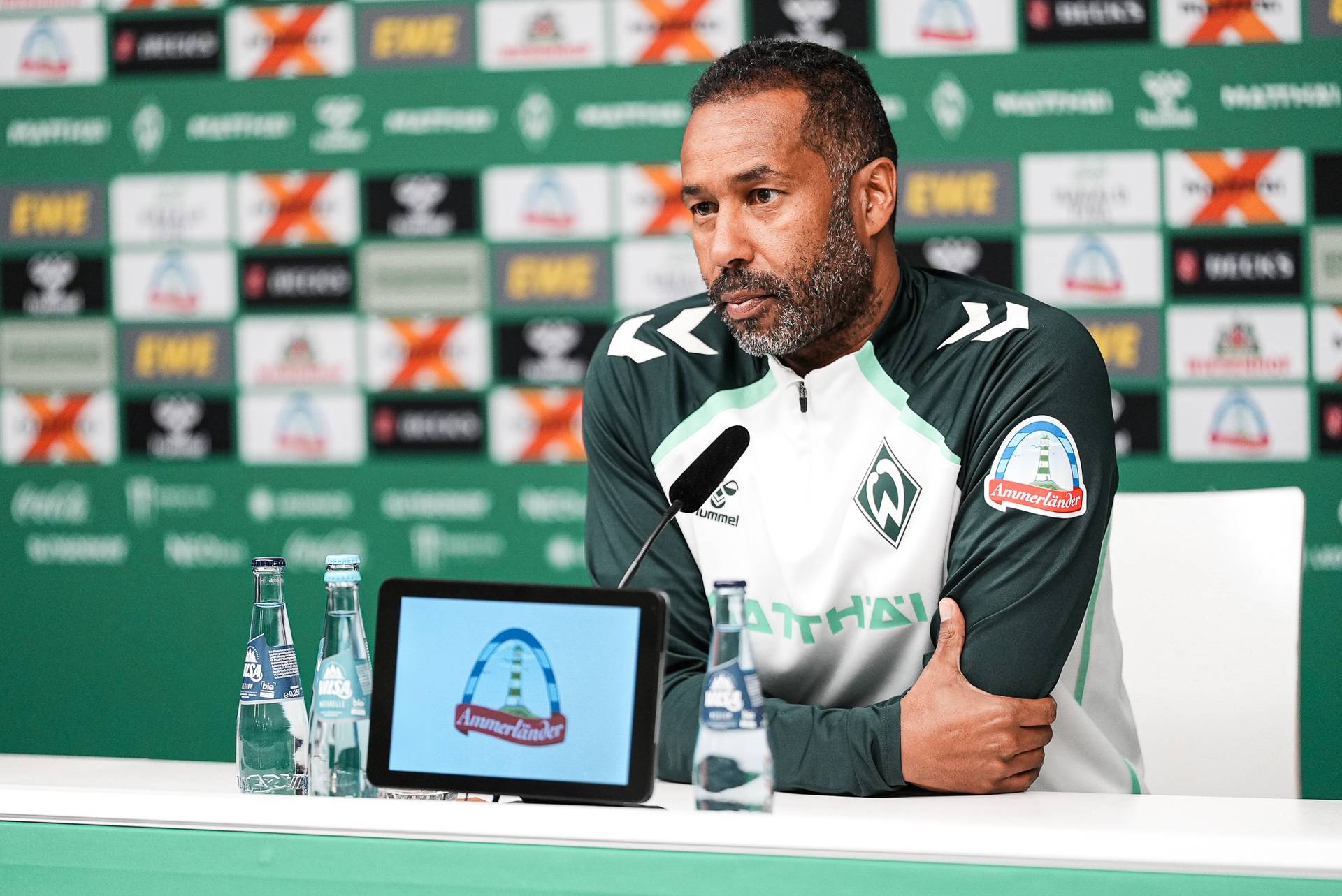 Ein Mann in einer grün-weißen Sportjacke spricht an einem Pressekonferenztisch, hinter ihm sind eine Leinwand und eine Markenhintergrundkulisse zu sehen.