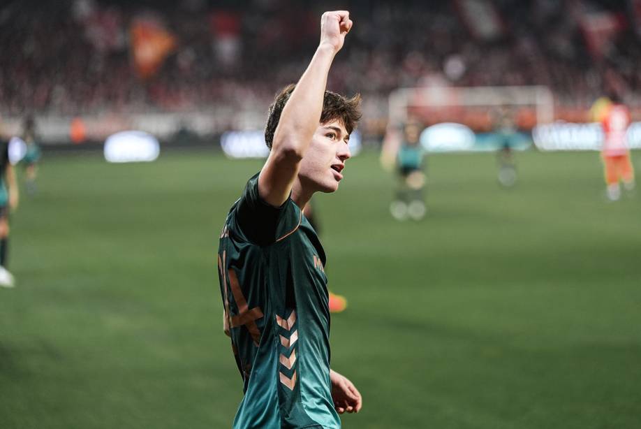 Fußballspieler in dunkelgrünem Trikot feiert mit erhobener Faust auf dem Spielfeld; verschwommene Menschenmenge im Hintergrund.