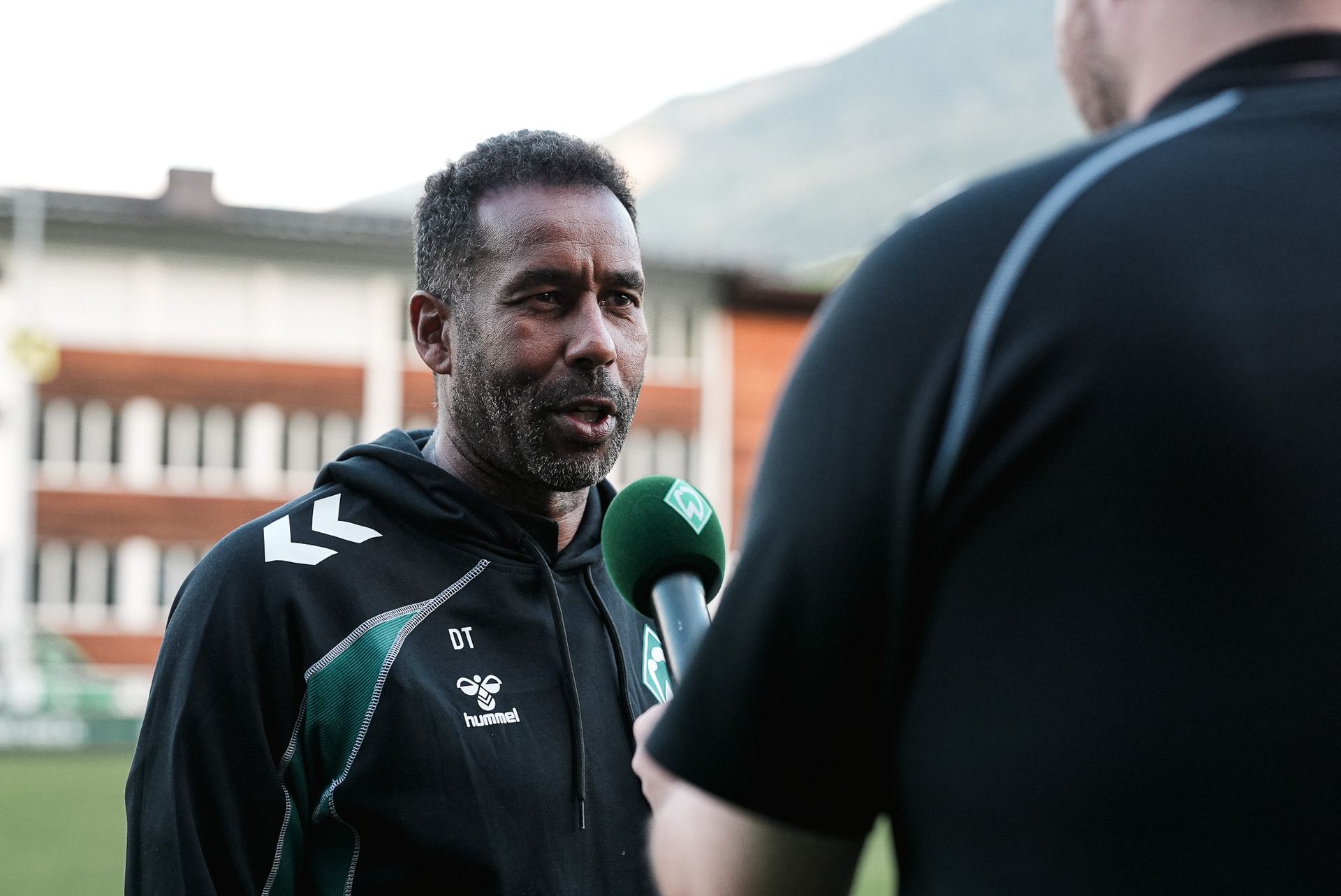 Ein Mann in einem Sport-Hoodie wird im Freien mit einem Mikrofon interviewt, im Hintergrund sind ein Gebäude und Berge zu sehen.