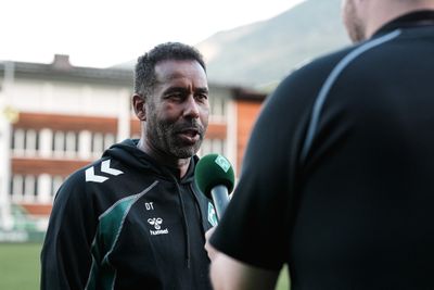 Ein Mann in einem Sport-Hoodie wird im Freien mit einem Mikrofon interviewt, im Hintergrund sind ein Gebäude und Berge zu sehen.