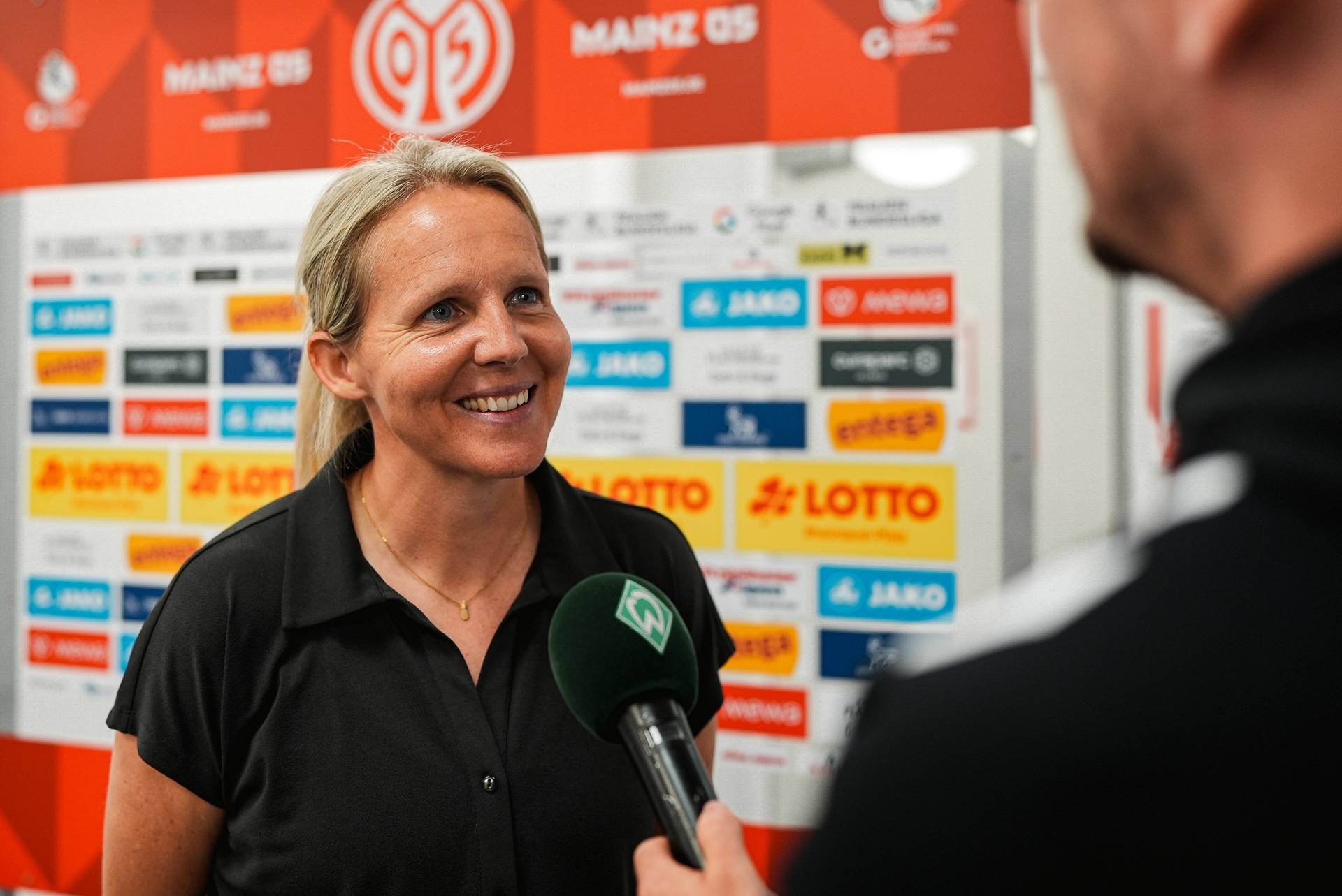 Eine Frau lächelt während eines Interviews mit einem Mikrofon vor einer Kulisse mit Mainz 05-Logos, die auf ein Sport-Sponsoring hinweist.