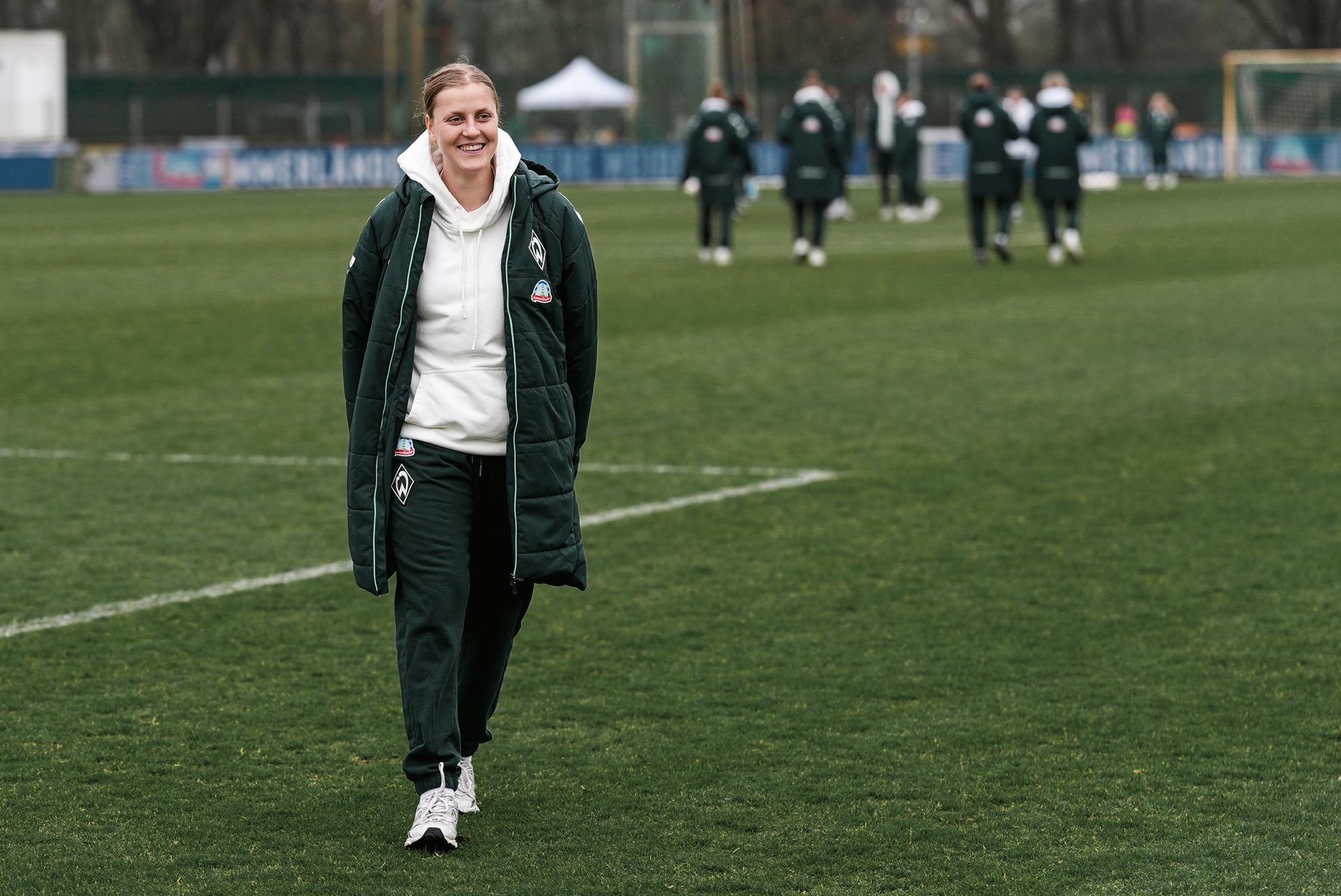 Eine Person in einer langen Jacke geht lächelnd über eine Wiese, während im Hintergrund andere Personen in ähnlicher Kleidung zu sehen sind.