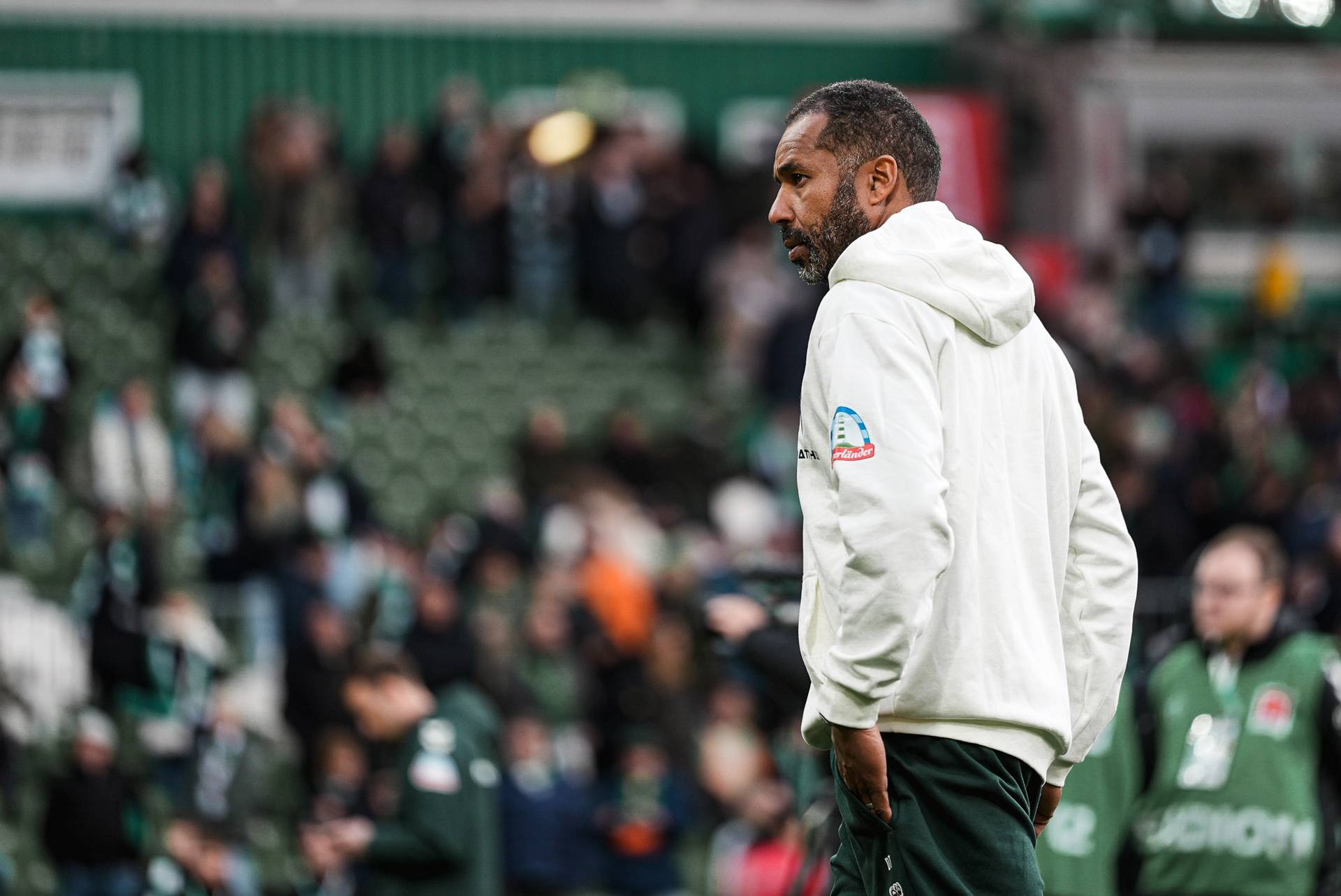 Ein Mann in einem weißen Kapuzenpulli steht auf einem Feld, im Hintergrund ist eine verschwommene Menschenmenge zu sehen; er wirkt nachdenklich.