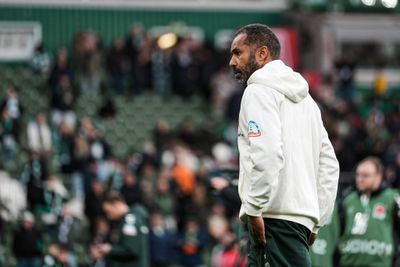 Ein Mann in einem weißen Kapuzenpulli steht auf einem Feld, im Hintergrund ist eine verschwommene Menschenmenge zu sehen; er wirkt nachdenklich.