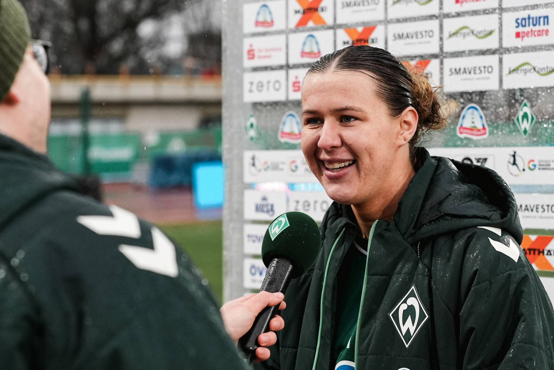 Ein lächelnder Fußballspieler in einer grünen Jacke wird interviewt und hält ein Mikrofon in der Hand. Im Hintergrund sind Sponsorenlogos zu sehen.