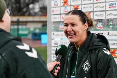 Ein lächelnder Fußballspieler in einer grünen Jacke wird interviewt und hält ein Mikrofon in der Hand. Im Hintergrund sind Sponsorenlogos zu sehen.