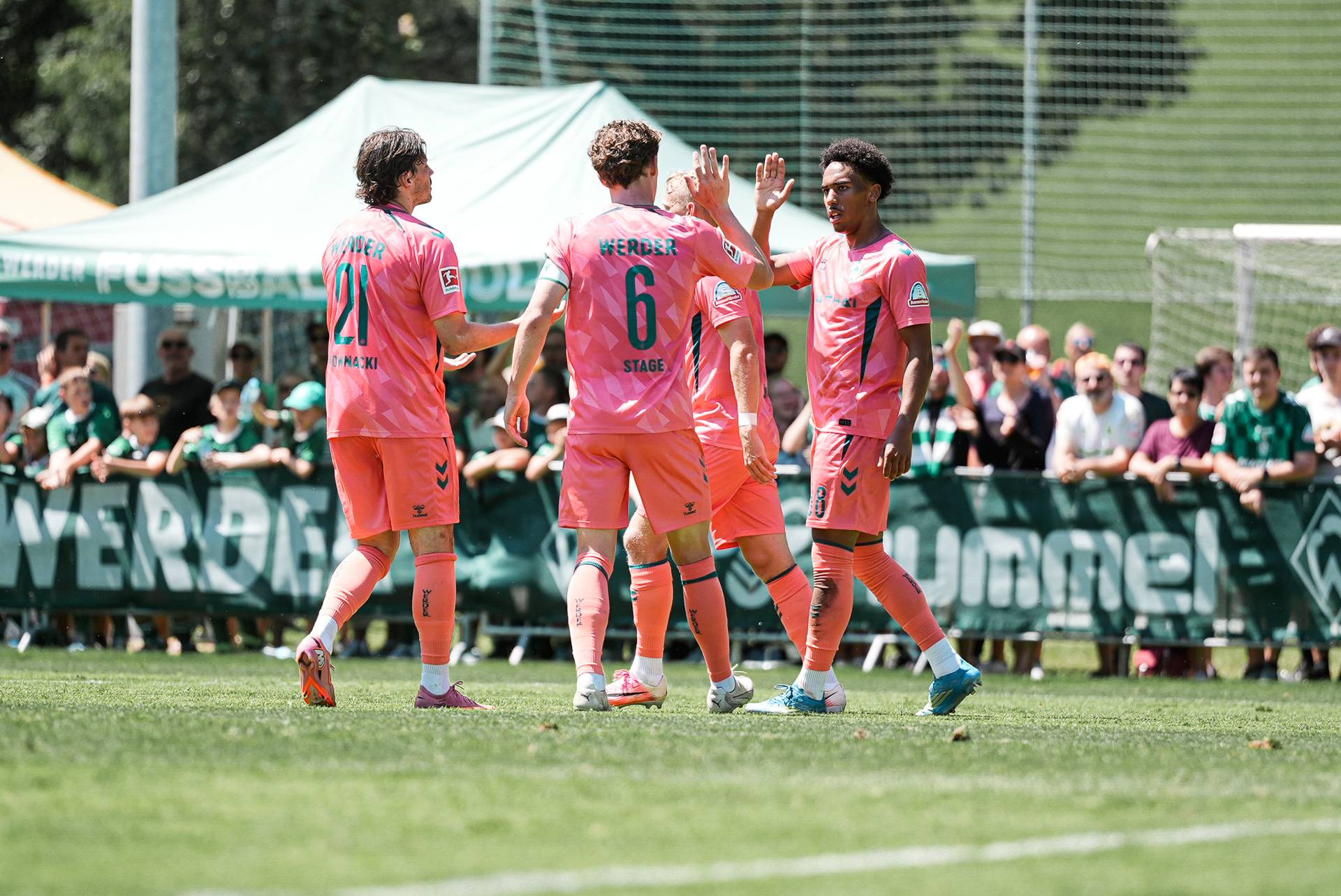 Fußballspieler in rosa Trikots klatschen sich auf dem Spielfeld ab, im Hintergrund ist eine Zuschauermenge zu sehen; sie tragen Trikots mit den Aufschriften „Werder“, „Stage“ und Rückennummern.
