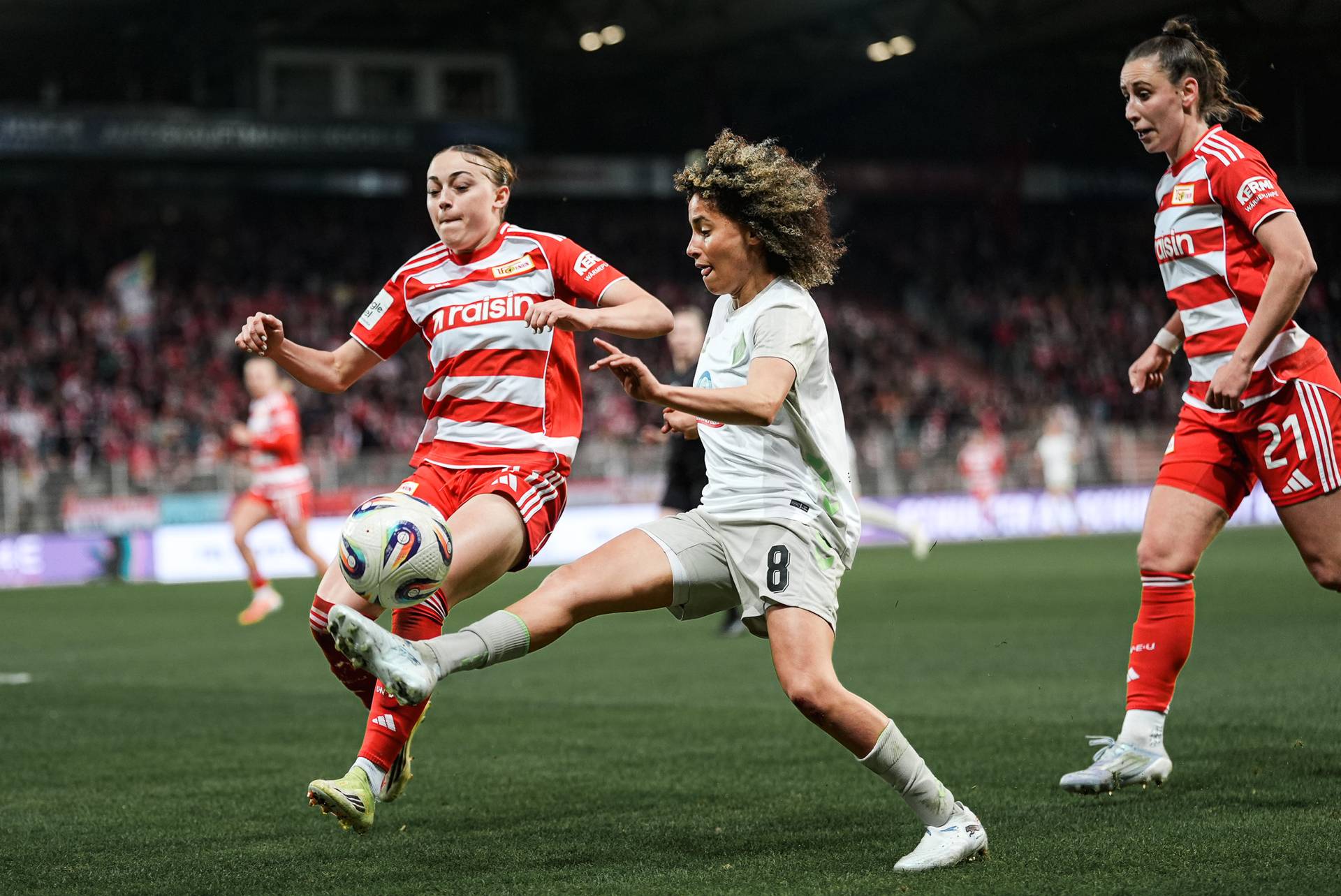 Zwei Fußballspielerinnen in rot gestreiften Trikots und einer in eine im weißen Trikot kämpfen auf dem Spielfeld um den Ball, während im Hintergrund eine gebannte Zuschauermenge steht.