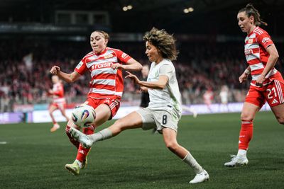 Zwei Fußballspielerinnen in rot gestreiften Trikots und einer in eine im weißen Trikot kämpfen auf dem Spielfeld um den Ball, während im Hintergrund eine gebannte Zuschauermenge steht.