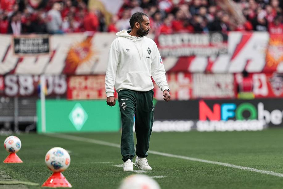 Ein Mann in einem weißen Kapuzenpulli und einer dunklen Hose steht auf einem Fußballplatz, umgeben von Pylonen. Im Hintergrund ist eine Menschenmenge mit roten Transparenten zu sehen.