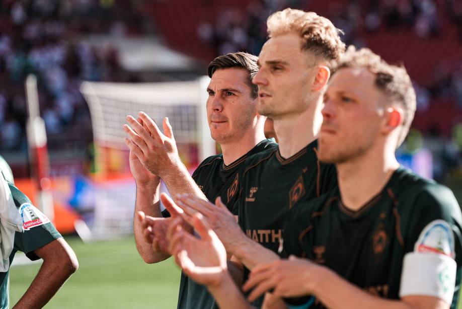 Drei Fußballspieler in grünen Trikots jubeln auf einem sonnigen Spielfeld, im Hintergrund sind verschwommene Zuschauer zu sehen.