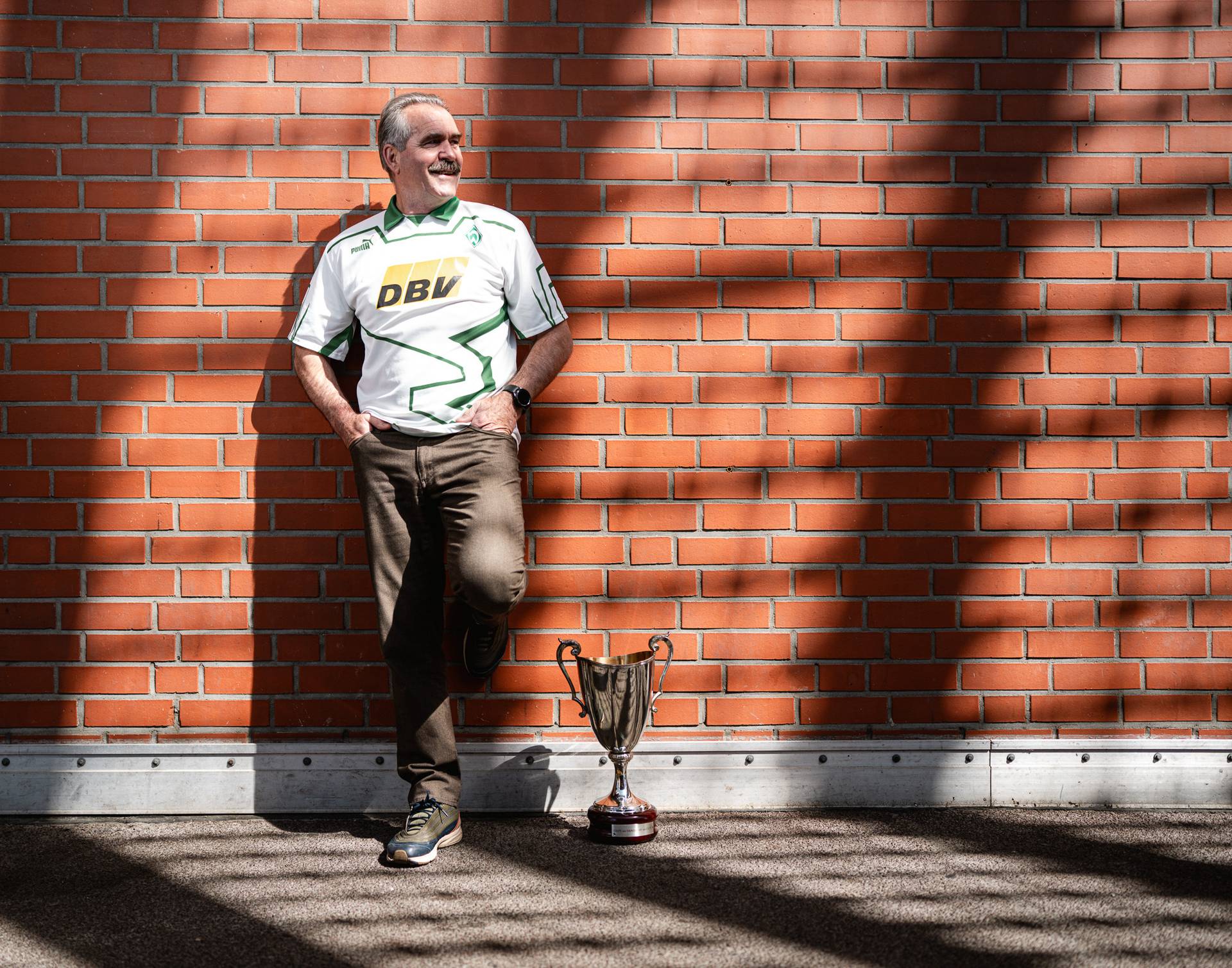 Ein Mann in einem weißen Sporttrikot lehnt lächelnd an einer Backsteinmauer, neben ihm steht eine Trophäe auf dem Boden. Schatten bedecken Teile der Mauer und des Bodens.