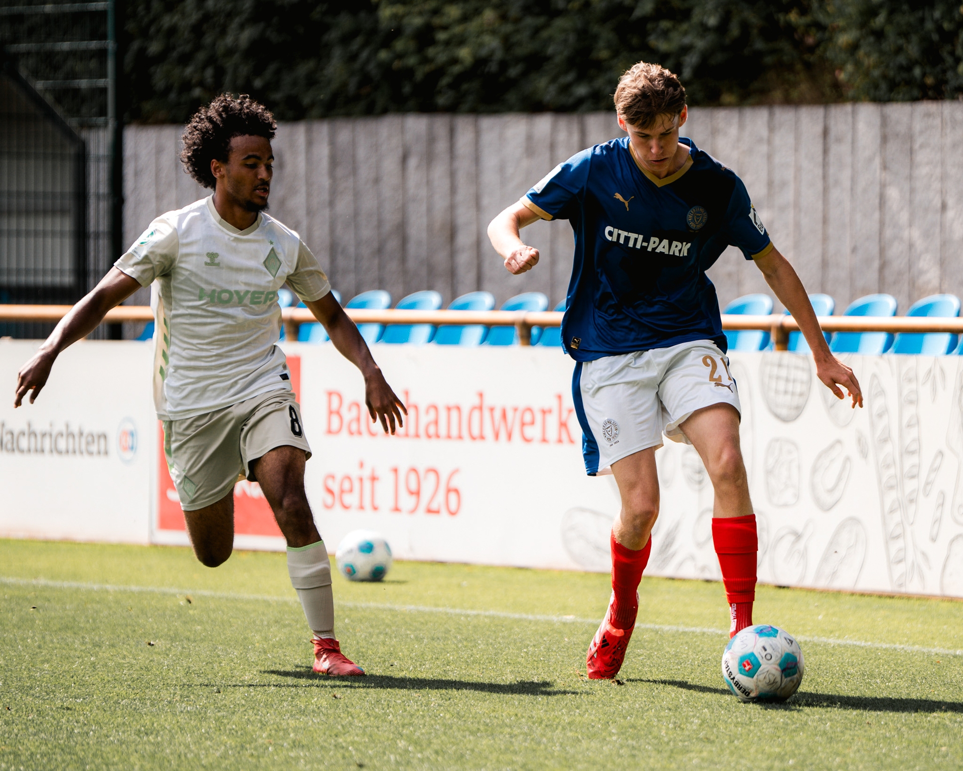 Zwei Fußballspieler kämpfen auf einem Rasenfeld um den Ball, wobei einer ein Werder Trikot und der andere ein Kiel Trikot trägt.