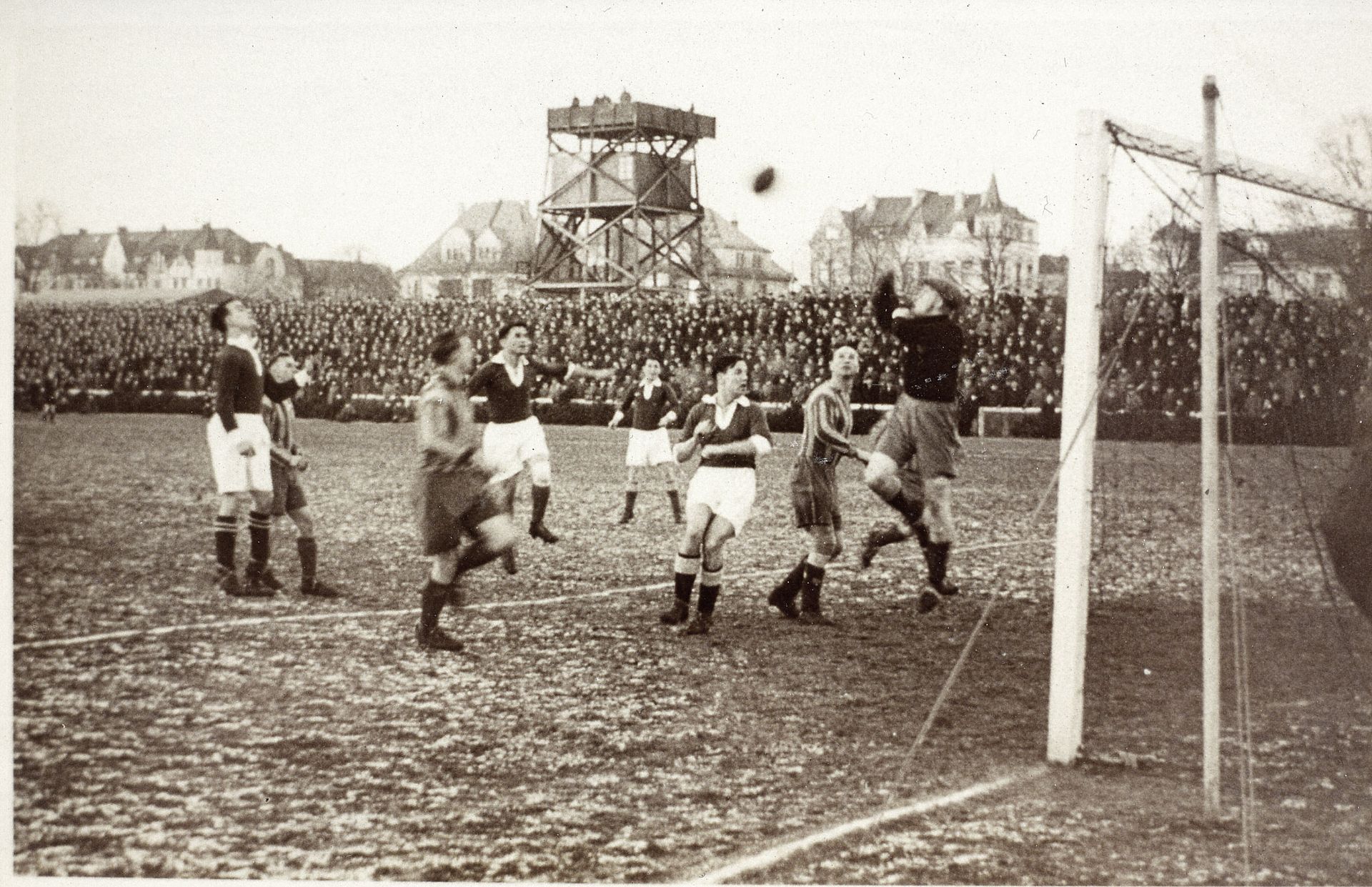 Ein Fußballspiel im Retro-Stil, bei dem die Spieler mitten im Spielgeschehen vor dem Tor zu sehen sind, umgeben von einer großen Zuschauermenge und alten Häusern im Hintergrund.