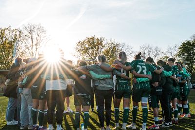 Eine Fußballmannschaft steht im Kreis auf einem Spielfeld, über das Sonnenlicht hereinströmt, im Hintergrund sind Bäume und Stadionbeleuchtung zu sehen.