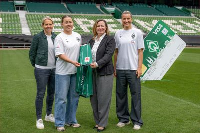 Vier Personen stehen auf einem Fußballplatz, im Hintergrund sind ein grünes Trikot und eine große Kreditkartenattrappe zu sehen.