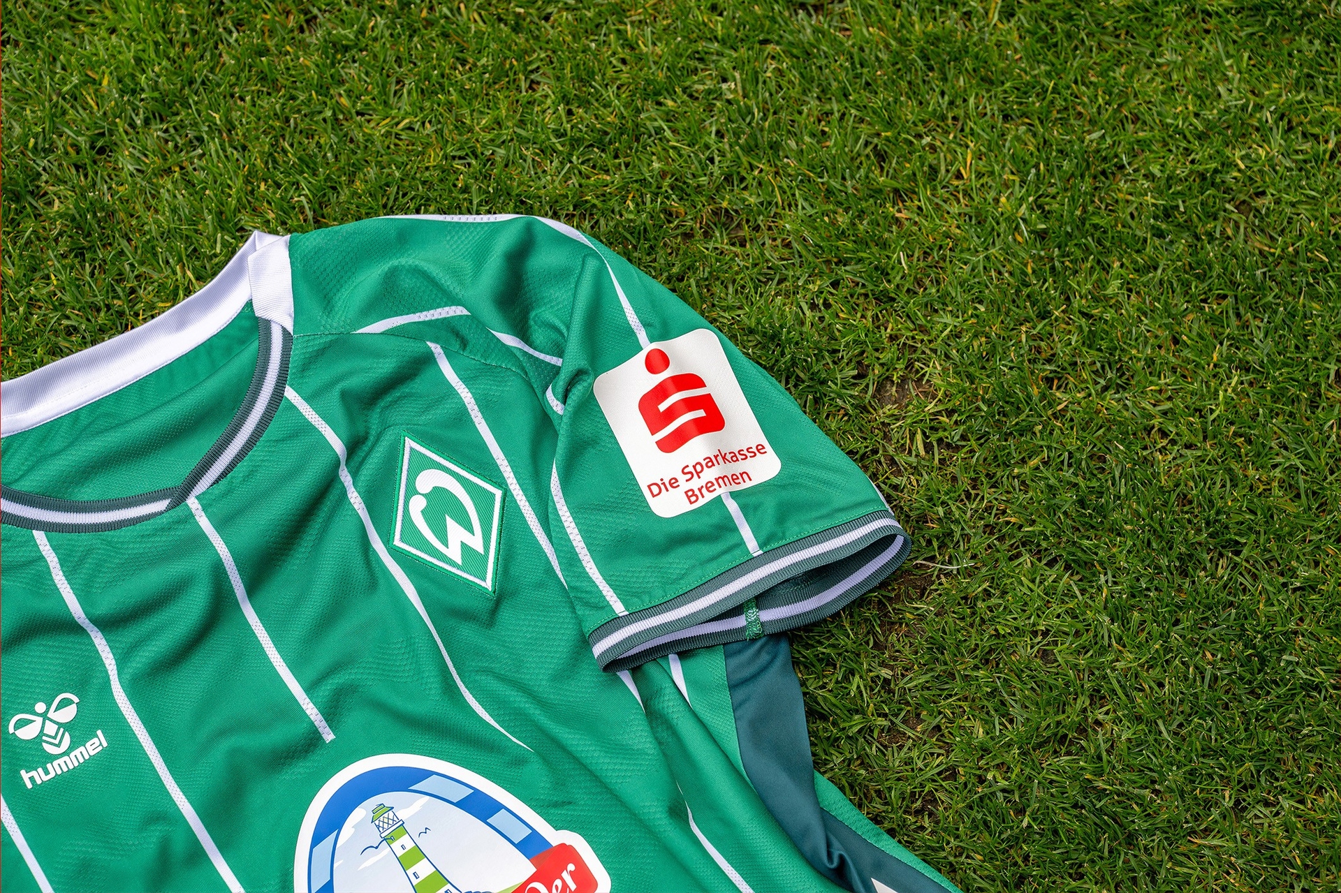 Grünes Fußballtrikot des SV Werder Bremen mit weißen Streifen auf Rasen, verziert mit den Logos von „hummel“, einem weißen „W“-Emblem und „Die Sparkasse Bremen“.