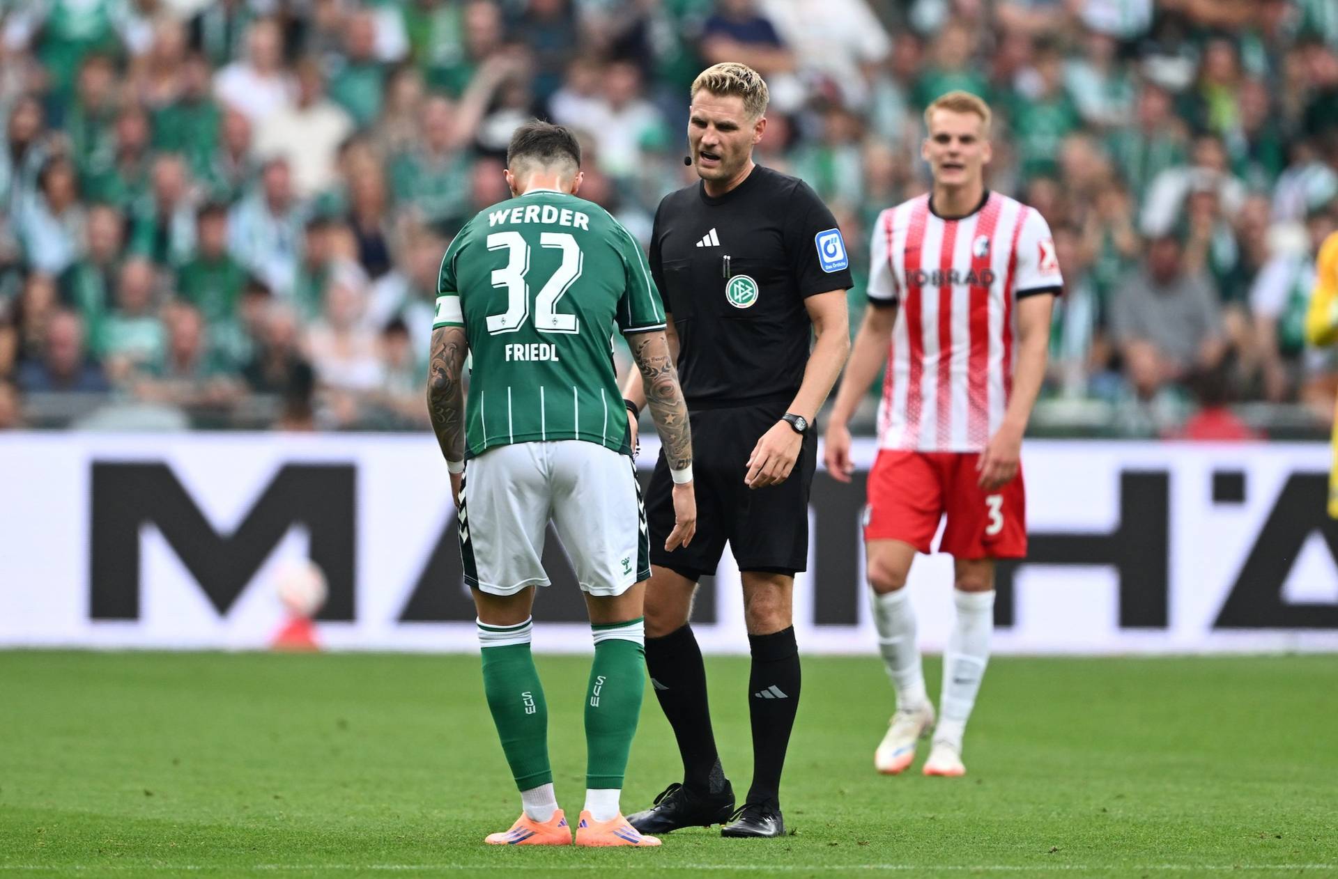 Fußballspieler in grünem Trikot steht einem Schiedsrichter gegenüber, während ein anderer Spieler in rot gestreiftem Trikot im Hintergrund auf einem Spielfeld steht.