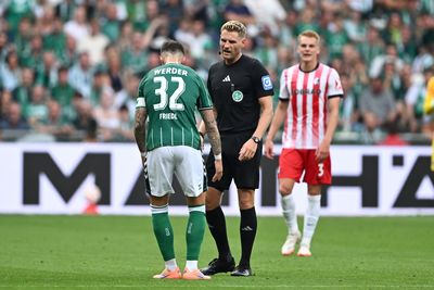 Fußballspieler in grünem Trikot steht einem Schiedsrichter gegenüber, während ein anderer Spieler in rot gestreiftem Trikot im Hintergrund auf einem Spielfeld steht.