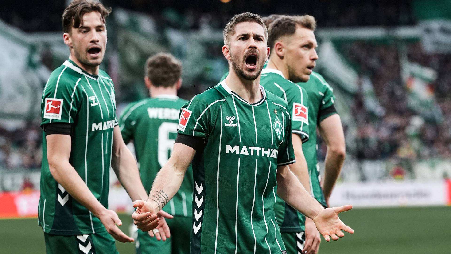 Fußballspieler in grünen Trikots reagieren leidenschaftlich auf dem Spielfeld, wobei ein zentraler Spieler während eines Spiels eindringlich gestikuliert.