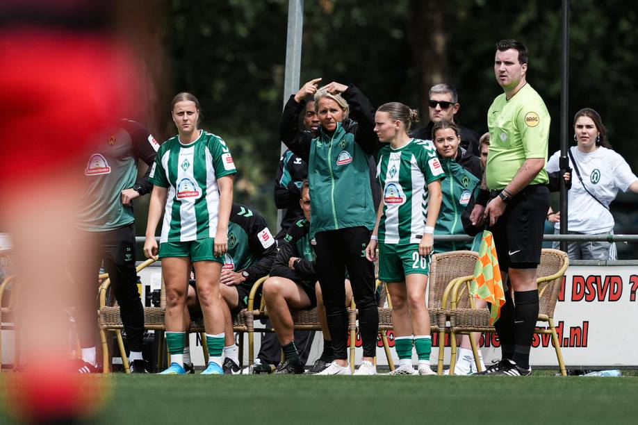 Während eines Spiels besprechen die Fußballspielerinnen und ihre Trainerin am Spielfeldrand die Taktik, während ein Schiedsrichter in der Nähe steht.