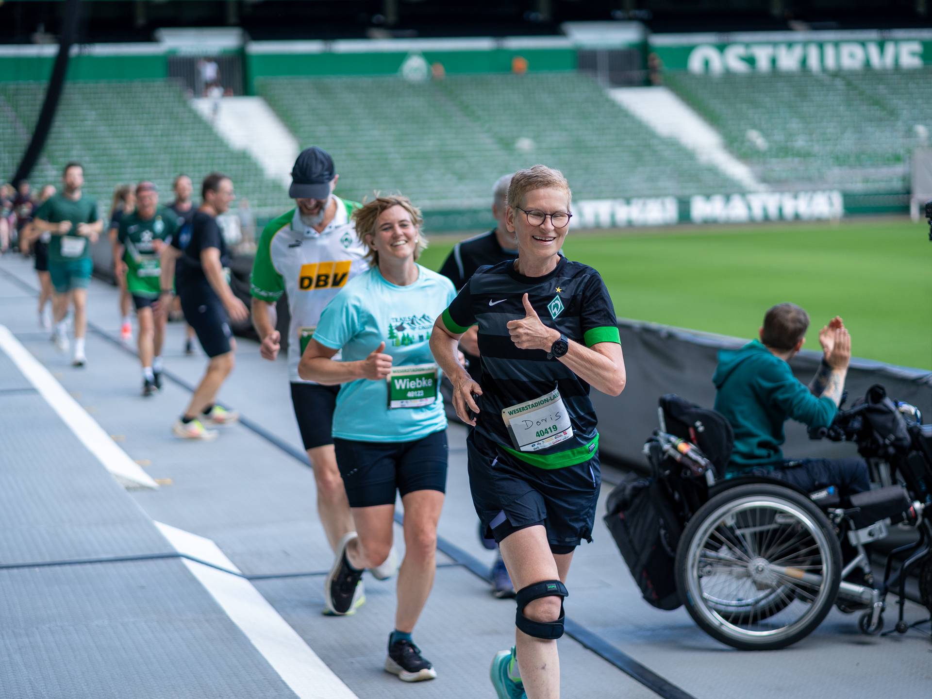 Läufer*innen, die an einem Marathon in einem Stadion teilnehmen, lächeln und zeigen den Daumen nach oben, während ein Zuschauer im Rollstuhl in der Nähe applaudiert.