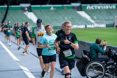 Läufer*innen, die an einem Marathon in einem Stadion teilnehmen, lächeln und zeigen den Daumen nach oben, während ein Zuschauer im Rollstuhl in der Nähe applaudiert.