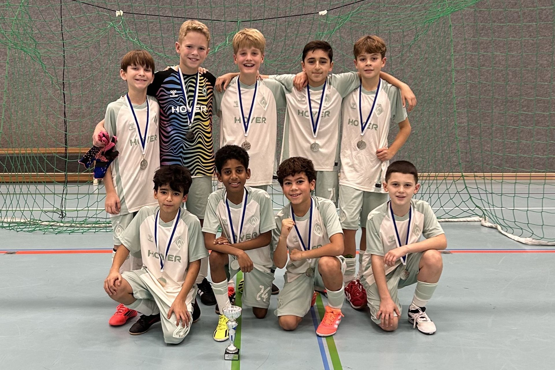 Neun Jungen einer Hallenfußballmannschaft in hellgrünen Trikots mit Medaillen, Torwart in buntem Trikot, posieren vor einem Tor um einen Pokal herum.