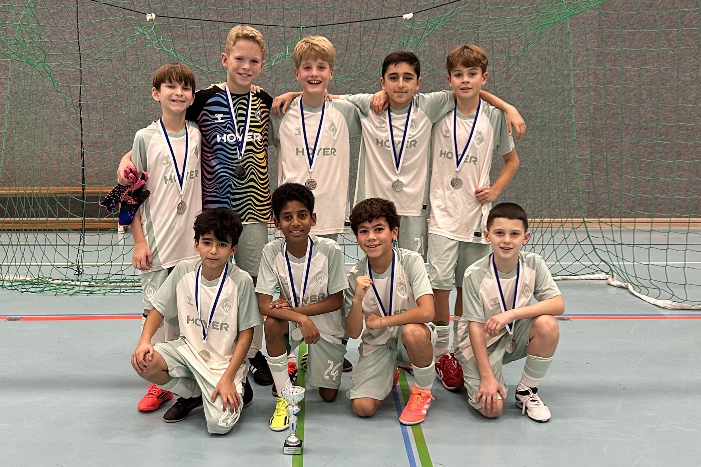 Neun Jungen einer Hallenfußballmannschaft in hellgrünen Trikots mit Medaillen, Torwart in buntem Trikot, posieren vor einem Tor um einen Pokal herum.