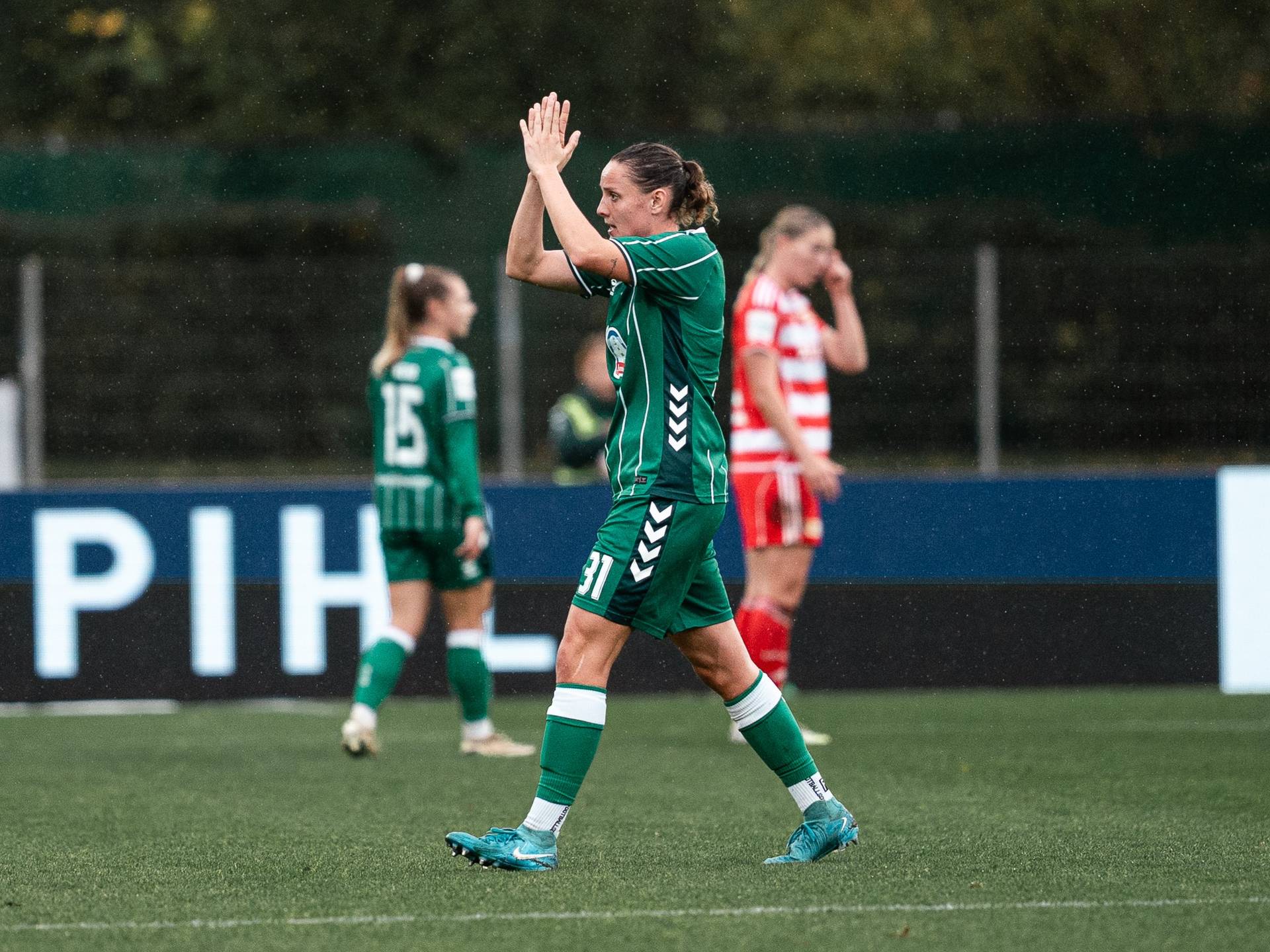 Verena Wieder applaudiert auf dem Spielfeld. Im Hintergrund sind Teamkolleginnen und eine Gegnerin zu sehen, eine davon in einem rot gestreiften Trikot.