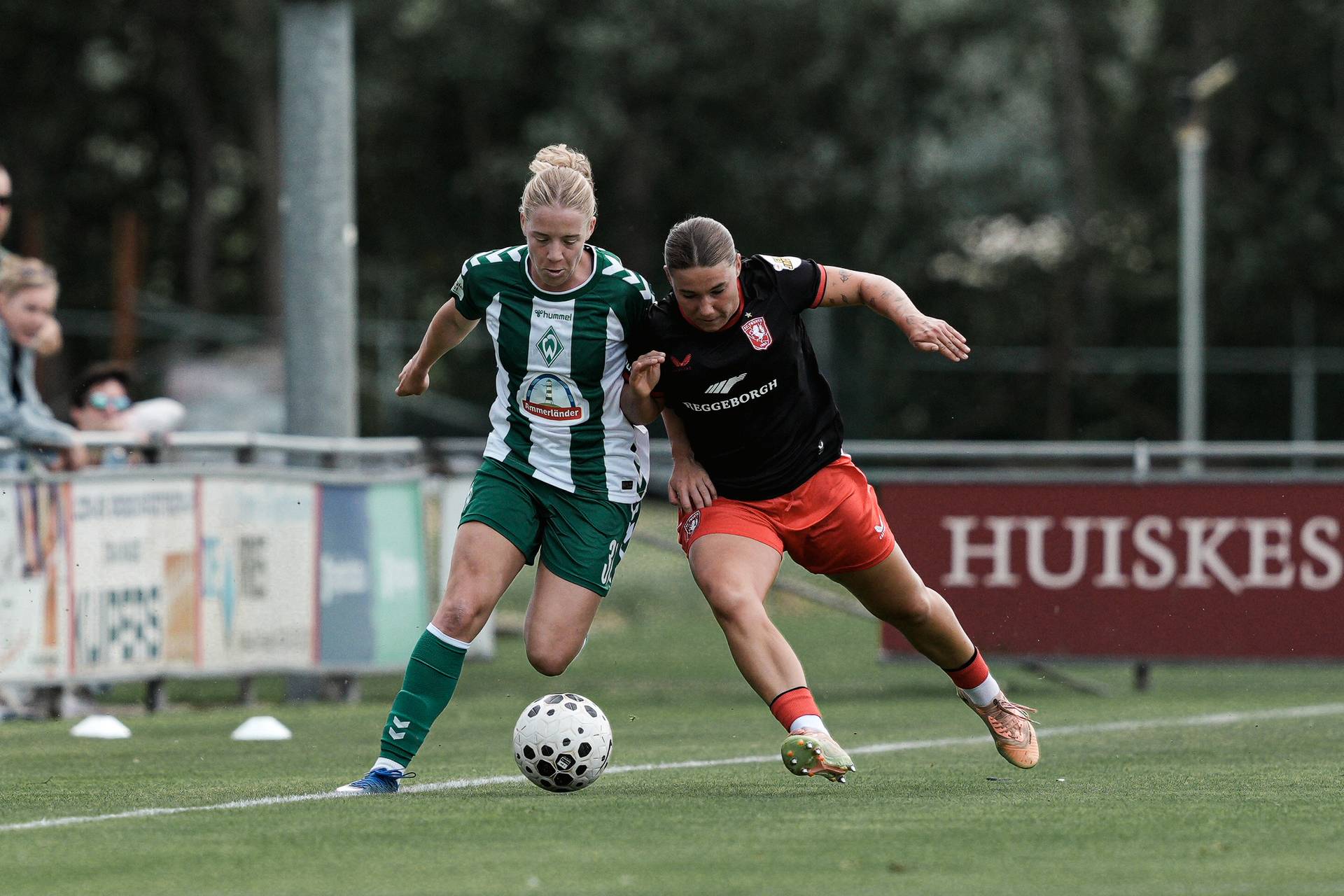 Zwei Fußballspielerinnen – eine in Grün und Weiß, die andere in Schwarz und Rot – kämpfen während eines Spiels auf einem Rasenfeld um den Ball.