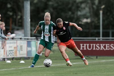 Zwei Fußballspielerinnen – eine in Grün und Weiß, die andere in Schwarz und Rot – kämpfen während eines Spiels auf einem Rasenfeld um den Ball.