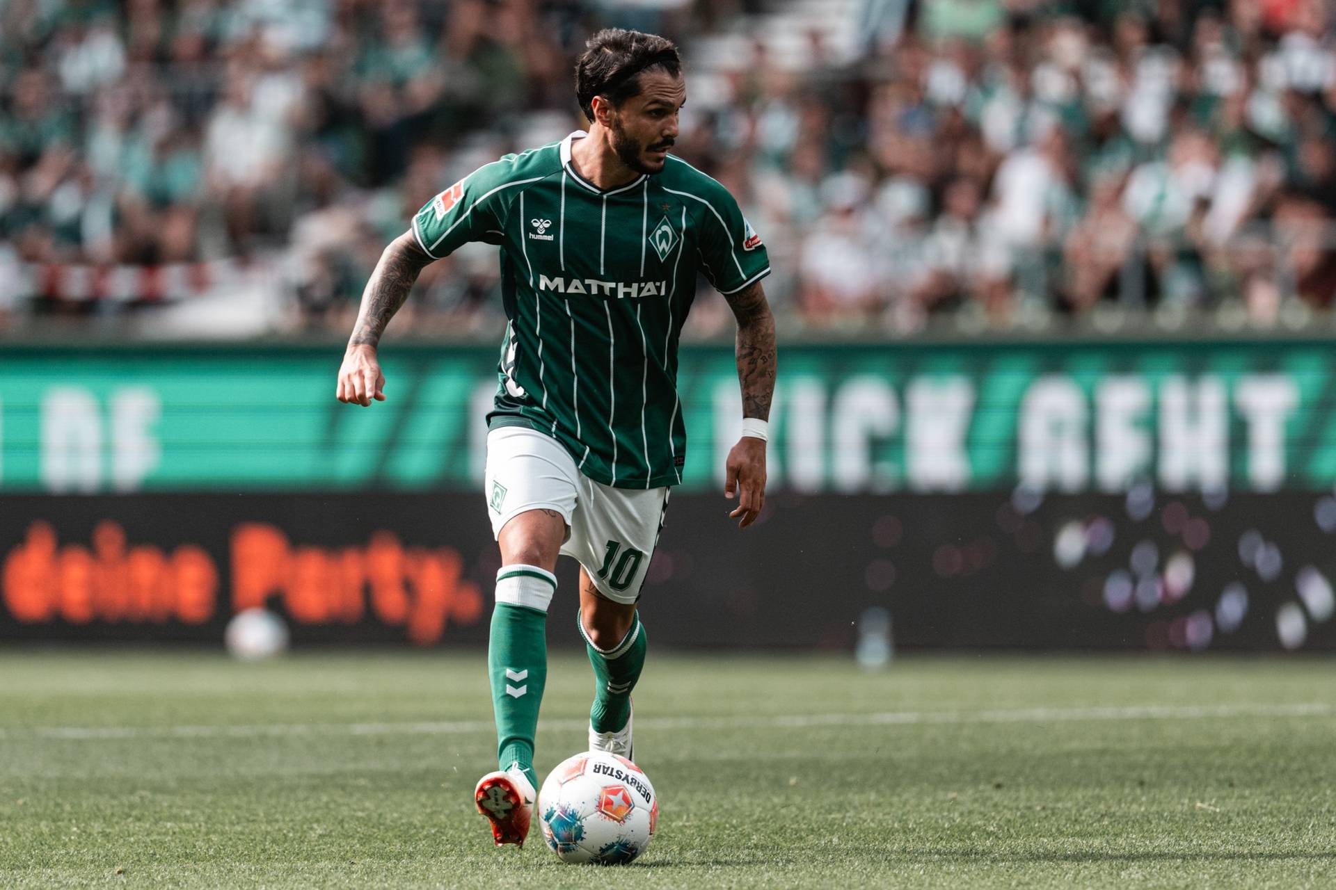 Leonardo Bittencourt dribbles with the ball at his feet. He's wearing the green Werder home kit with thin white stripes, as well as white shorts and green socks.