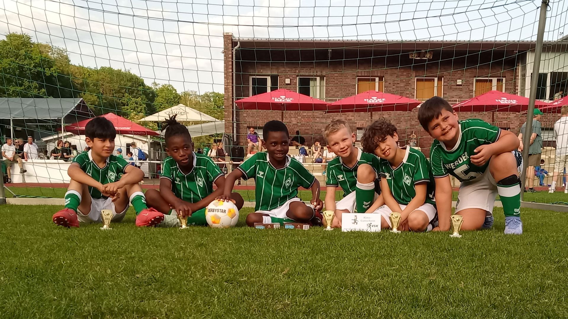 Sechs junge Fußballspieler in grünen Trikots posieren mit Pokalen auf einem Spielfeld und lächeln vor einem Tor.