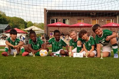 Sechs junge Fußballspieler in grünen Trikots posieren mit Pokalen auf einem Spielfeld und lächeln vor einem Tor.