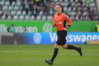 Ein Fußballschiedsrichter in orangefarbener Uniform rennt über einen grünen Rasen, im Hintergrund sind eine verschwommene Menschenmenge und das Volkswagen-Logo zu sehen.