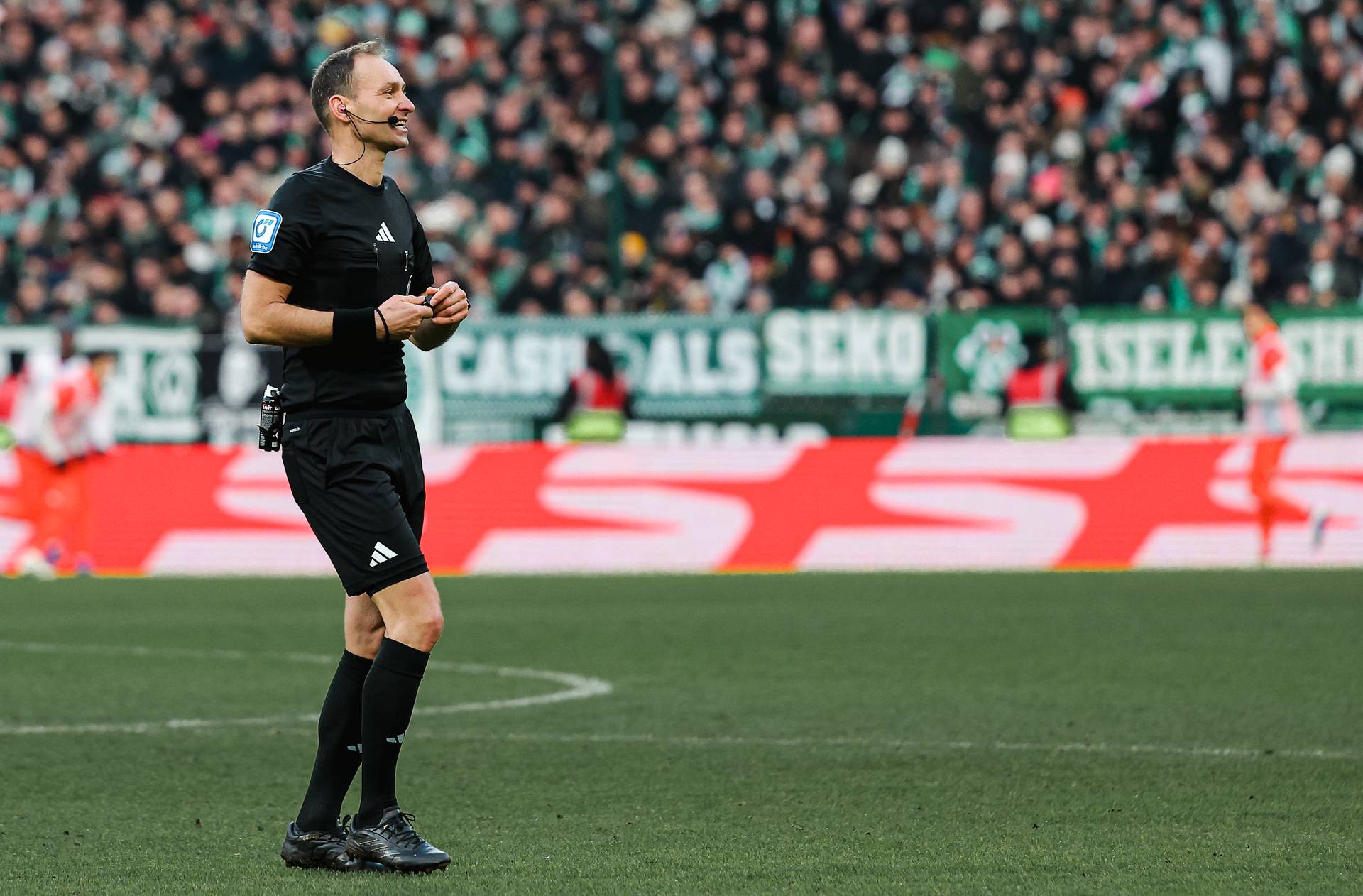 Ein Fußballschiedsrichter in schwarzer Uniform steht lächelnd auf dem Spielfeld, im Hintergrund ein vollbesetztes Stadion und verschwommene Spieler.