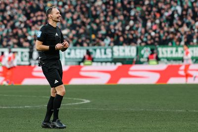 Ein Fußballschiedsrichter in schwarzer Uniform steht lächelnd auf dem Spielfeld, im Hintergrund ein vollbesetztes Stadion und verschwommene Spieler.
