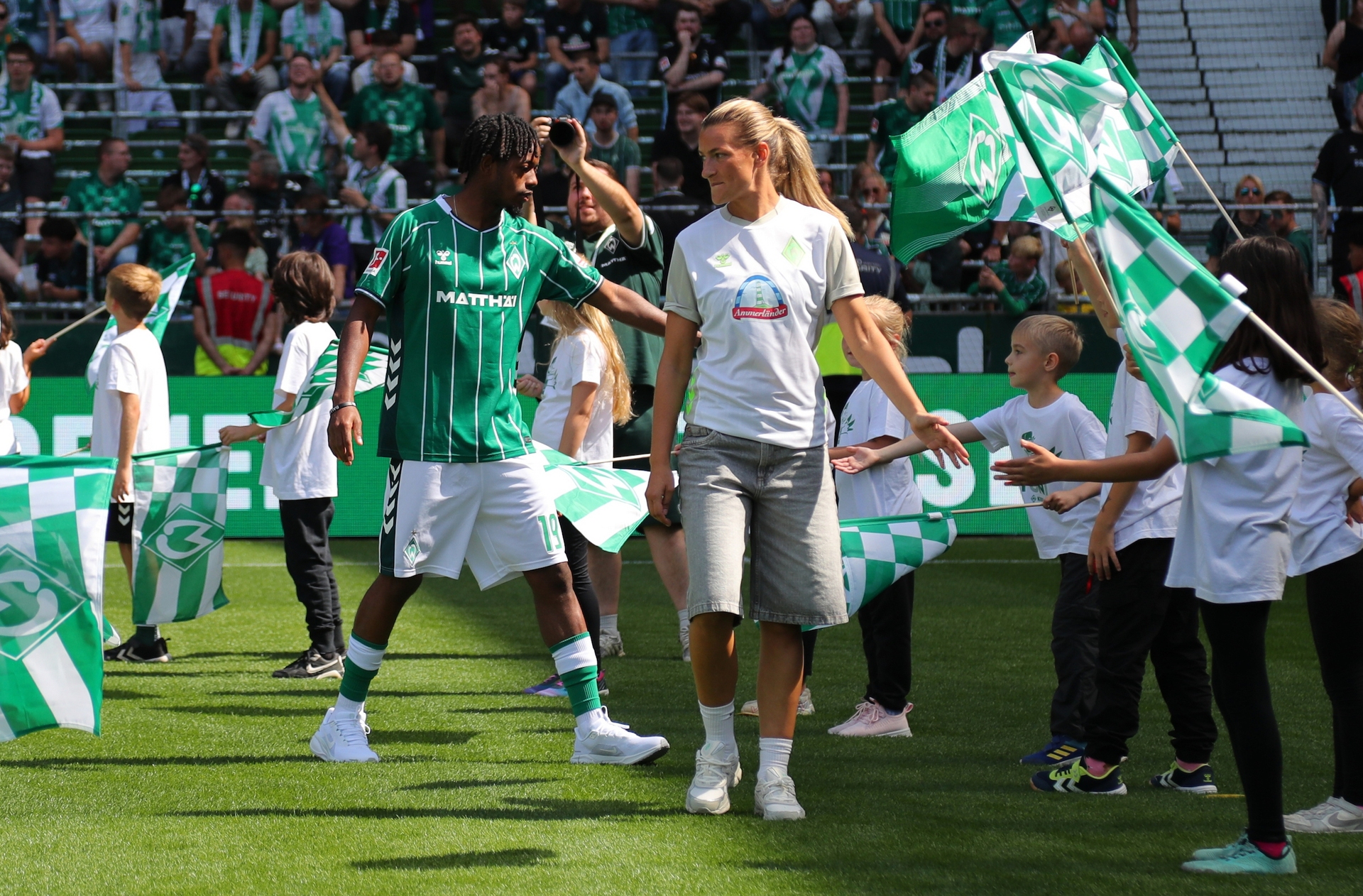 Samuel Mbangula und Medina Desic laufen ein und klatschen dabei Kinder mit Werder-Fahnen in der Hand ab,