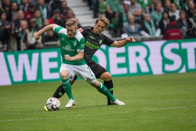 Zwei Fußballspieler in Aktion während eines Spiels. Der eine im grünen Trikot kontrolliert den Ball, während der andere im schwarzen Trikot versucht, ihn ihm abzunehmen.