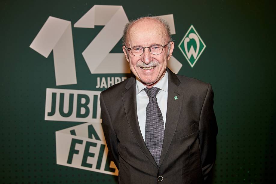 Willi Lemke lächekt im Anzug. Er steht vor einer Wand, auf der das 125-Jahre Werder-Logo abgebildet ist.