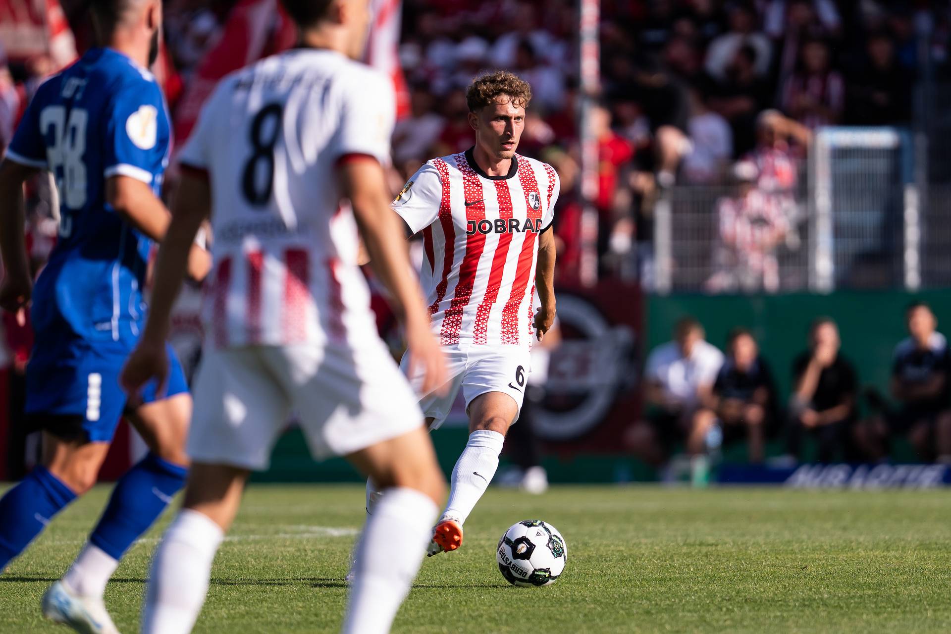 Ein Fußballspieler in einem rot-weiß gestreiften Trikot dribbelt während eines Spiels auf einem Feld mit dem Ball, umgeben von anderen Spielern.