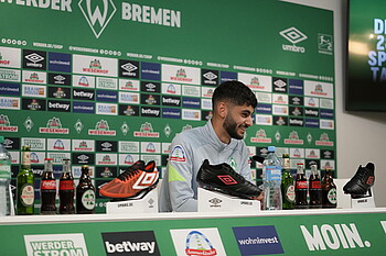 null Eren Dinkci sitzt lächelnd auf am Pult im PK-Raum.