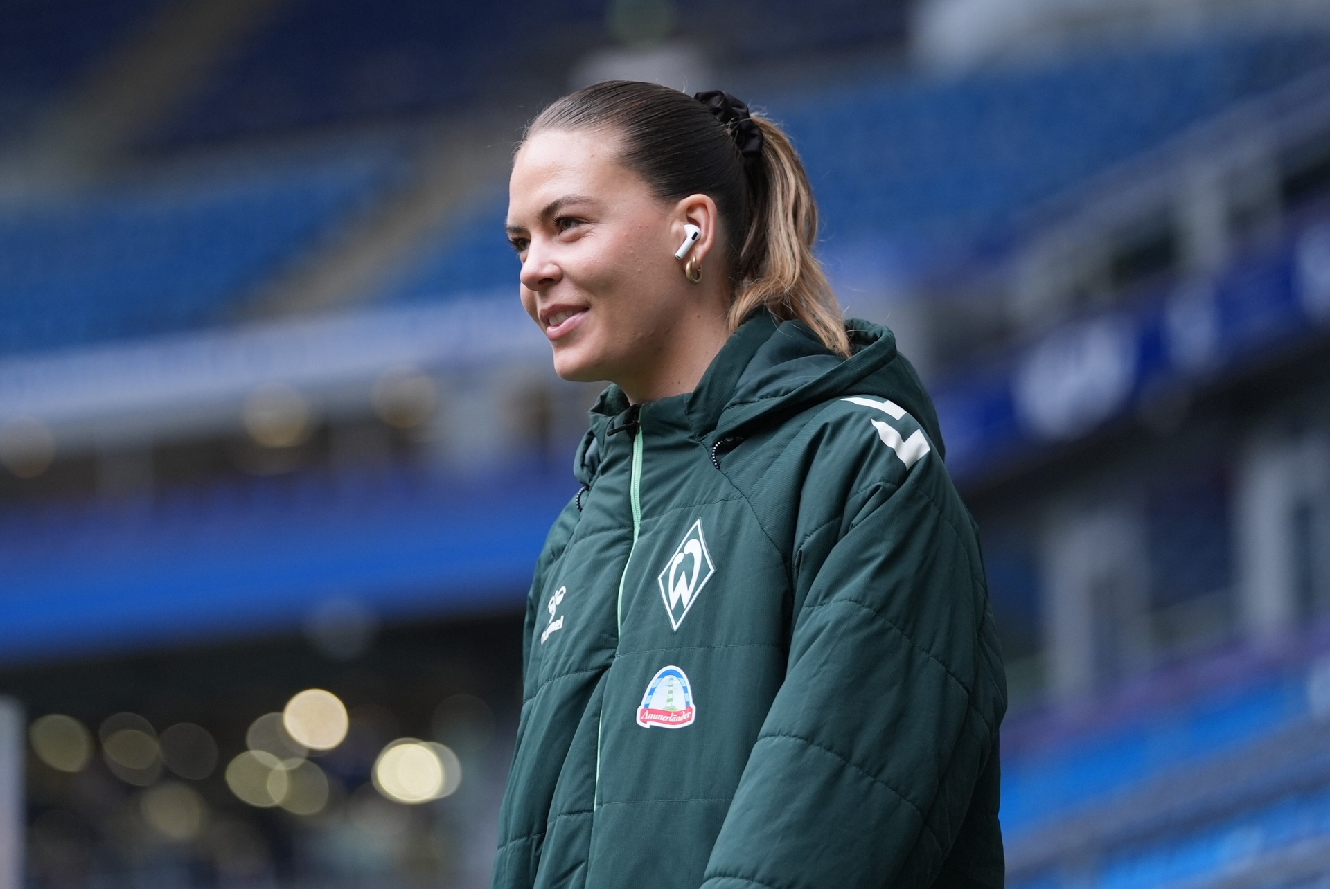 Juliane Wirtz im Profil. Sie trägt eine dunkelgrüne Jacke und hat weiße Kopfhörer in den Ohren. Im Hintergrund sind blaue Tribünen zu sehen.