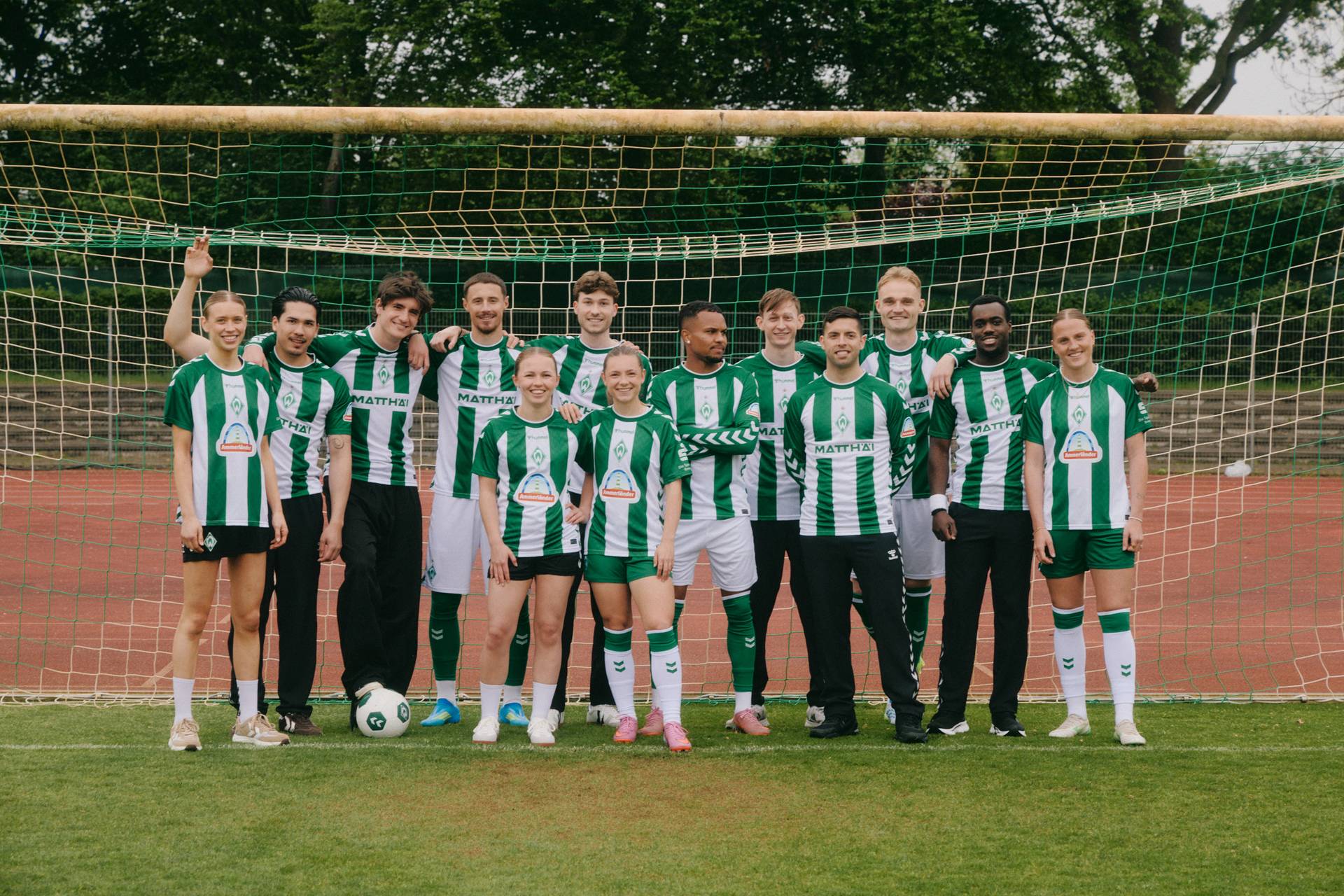 Männer und Frauen posieren in grün-weiß gestreiften Trikots in einem Fußballtor. Im Hintergrund sind Bäume zu sehen.