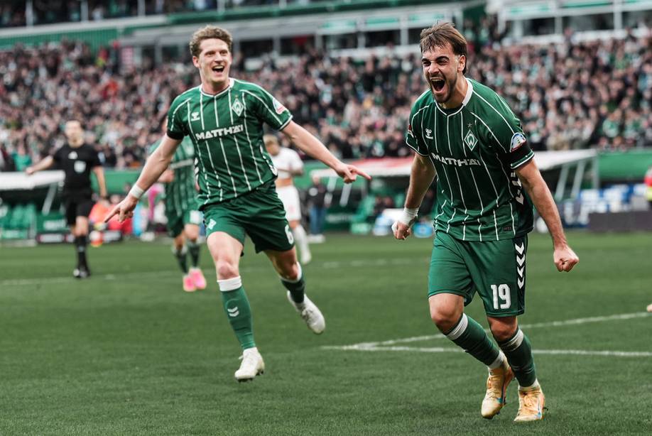Zwei Fußballspieler in grün gestreiften Trikots feiern ein Tor auf dem Spielfeld, im Hintergrund jubelt die Menge.