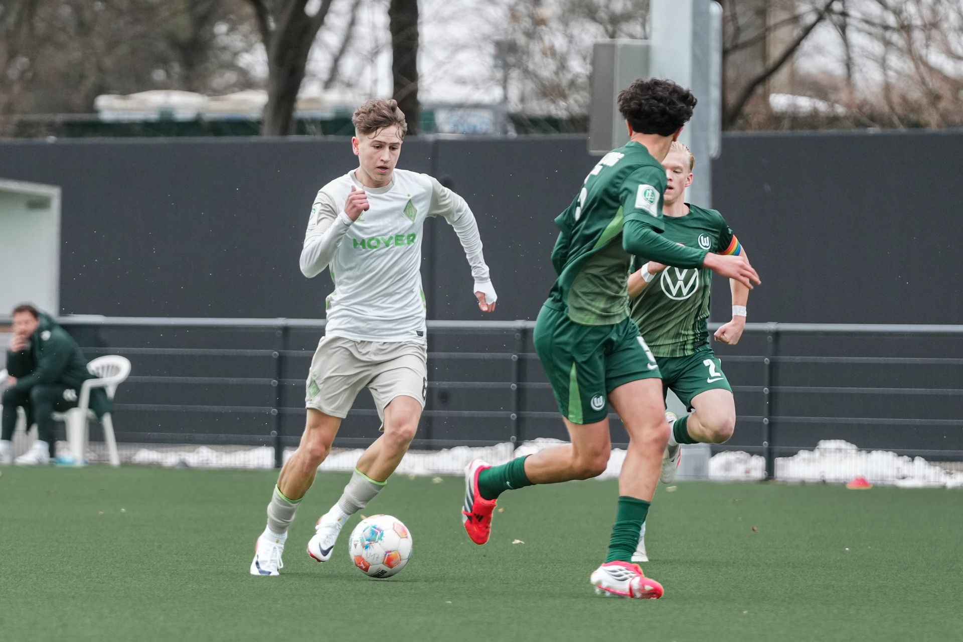 Fußballspieler in grün-weißen Trikots kämpfen auf einem Spielfeld um den Ball, im Hintergrund sitzen einige Zuschauer.