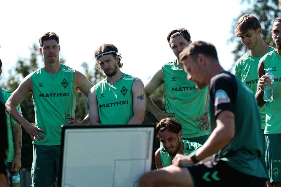 Eine Fußballmannschaft hört einem Trainer während einer Trainingseinheit aufmerksam zu und hat sich an einem sonnigen Tag um eine Taktiktafel versammelt.