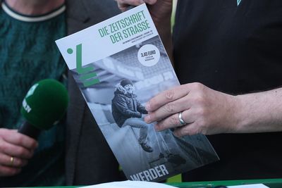 Eine Person, die eine Zeitschrift mit dem Titel „Die Zeitschrift der Straße“ in der Hand hält und neben jemandem mit einem grünen Mikrofon steht.